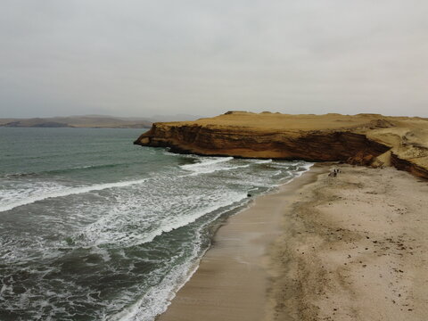 Aerial view of the rugged coastline of Paracas National Reserve with sandy cliffs, crashing waves, and a sandy beach under an overcast sky in Paracas, Ica, Peru.