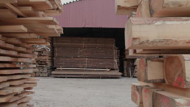 Large stacks of rough-sawn timber and wooden boards stored outdoors beside a covered industrial building at a lumber yard, showing organized wood inventory