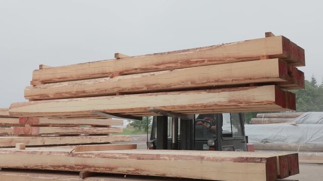 Forklift operator moving large rough-sawn timber beams in an outdoor lumber yard, illustrating heavy material handling, storage, and wood industry logistics
