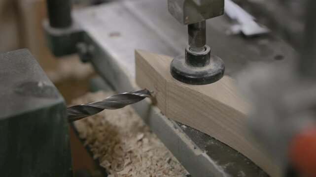 Close-up of a drill press boring into a wooden beam secured on a workbench, with wood shavings and sawdust visible around the drilling area