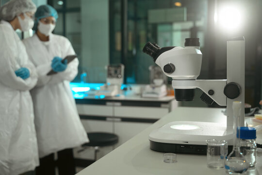 Stereo Microscope on Lab Bench with Scientists in White Coats and Masks Discussing Research in Modern Laboratory