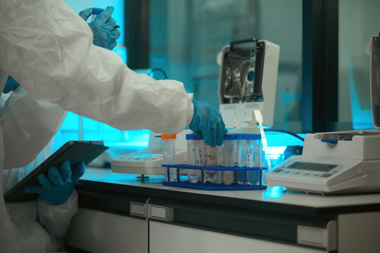 Close up of Pharmaceutical Lab Scientist in Protective Suit Handling Test Tubes with Digital Tablet for Quality Control Research