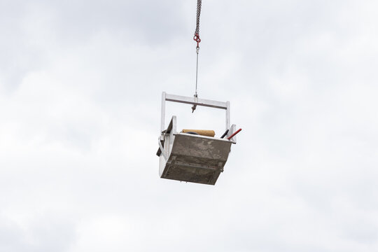 A container filled with roofing tools and equipment is suspended from an industrial crane.  Roof repair work, maintenance, transport, outdoors during the day, cloudy sky.