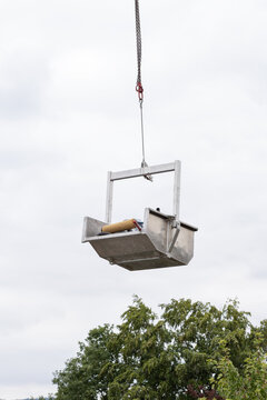 A container filled with roofing tools and equipment is suspended from an industrial crane.  Roof repair work, maintenance, transport, outdoors during the day, cloudy sky.