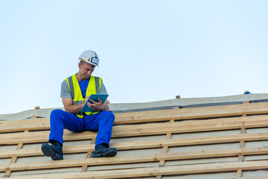 Construction worker wearing protective hard hat and high visibility vest inspecting building plans and taking notes on a new house roof framing against a clear sky