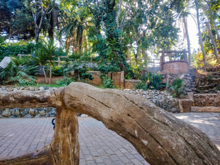 Rustic wooden park fence carved like a tree trunk stands prominently in the foreground overlooking lush green garden paths under bright natural afternoon sunny light © Graphixa