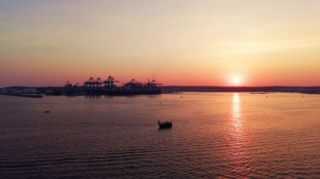 Sunset over the Mediterranean Sea with a silhouetted Maltese luzzu and Malta Freeport visible in the distance, captured in a wide-angle drone shot with slow forward movement over calm water.