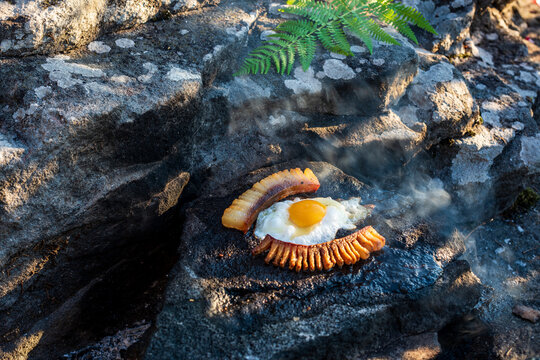 Outdoor Cooking of Fried Egg and Bacon on Hot Rock. Fried egg and bacon cooked directly on a hot stone outdoors, surrounded by natural rocks and green fern, creating a rustic outdoor cooking scene.