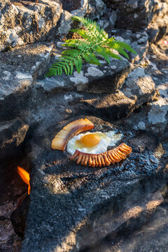 Outdoor Cooking of Fried Egg and Bacon on Hot Rock. Fried egg and bacon cooked directly on a hot stone outdoors, surrounded by natural rocks and green fern, creating a rustic outdoor cooking scene.