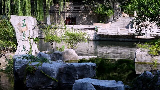 A tranquil outdoor garden scene featuring a stone pool, inscribed rock, and lush green plants and trees.