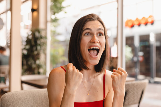 Ecstatic Caucasian woman celebrating success holding raised fists shouting looking away wearing orange top, horizontal portrait of happy winner girl reacting to good news in bright city cafe setting