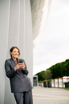 Woman checks phone by modern building on cloudy day near green trees