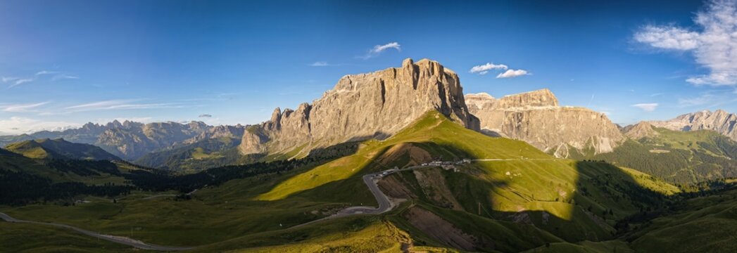 Aerial view of Sella Pass with its winding mountain road, lush green alpine meadows, and dramatic limestone peaks under a clear blue sky in Selva, Trentino-Alto Adige, Italy.