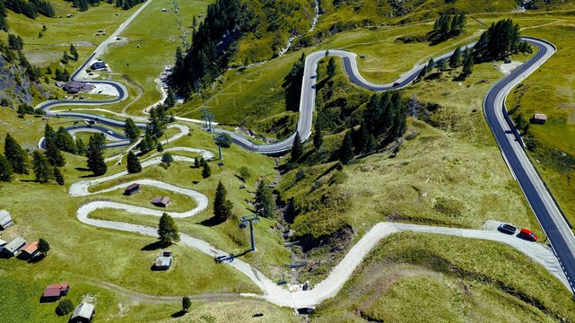 Aerial view of the winding mountain road at Passo Fedaia with lush green slopes, alpine meadows, and scattered wooden huts in Rocca Pietore, Veneto, Italy.
