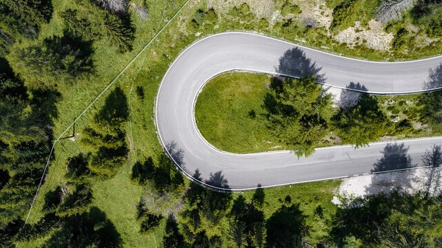 Aerial view of a sharp hairpin turn on a mountain road winding through the lush green forests of the Falzarego Pass in Cortina d'Ampezzo, Veneto, Italy.