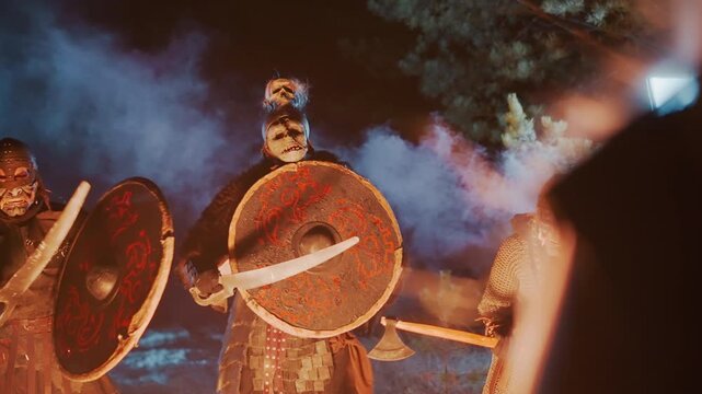 Three armored fighters move forward through drifting blue smoke at night, shields held upright and wooden polearms ready for combat, with warm firelight below and cool backlight shaping tension