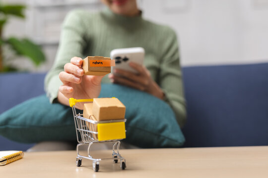 Woman using smartphone while holding return parcel at home, representing online shopping, product return, refund service, e-commerce delivery, customer support and retail purchase.
