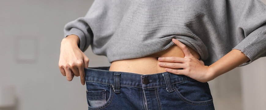 Unrecognizable young adult woman shows proud emotional success of weight loss diet holding large oversized denim jeans away from thin body and waist inside home living room