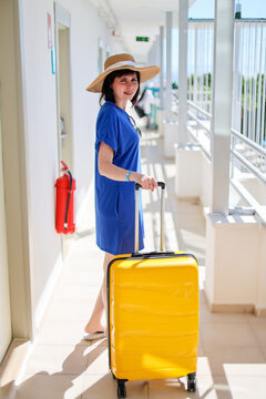 Happy woman tourist blue dress and sun hat with bright yellow suitcase at sunny hotel near her room enjoying summer vacation and travel.