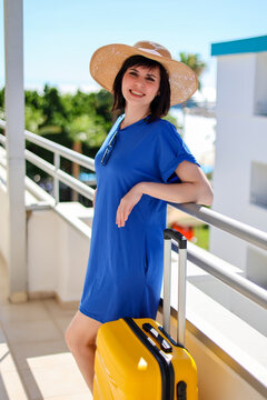 Happy woman tourist in blue dress and sun hat with bright yellow suitcase at sunny hotel near her room enjoying summer vacation and travel.