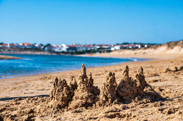 Close-up of textured drip-sand castle towers built on a sandy beach, with calm blue water and a whitewashed coastal town blurred in the background under a clear sky. © ricardo rocha