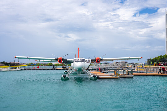 A propeller seaplane floats on clear turquoise water docked beside a terminal pier in Maldives under a cloudy summer sky.
