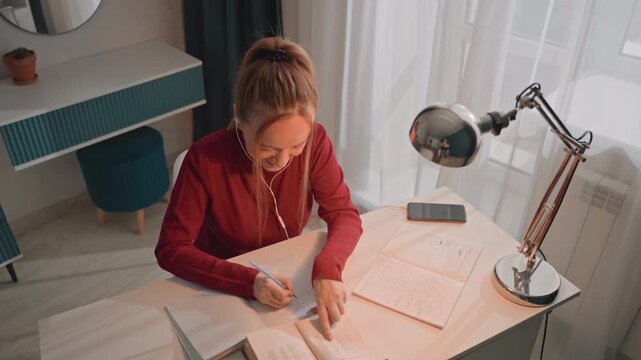 Young woman in red long sleeve top studies at desk writing. Desk features lined paper smartphone on charger metallic. Scene remains consistent as woman focuses on study materials