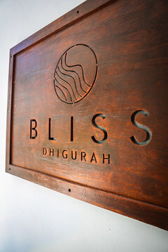 A brown wooden carved signboard displaying Bliss Dhigurah with a circular wave logo mounted on a plain white wall at a tropical resort in Maldives.