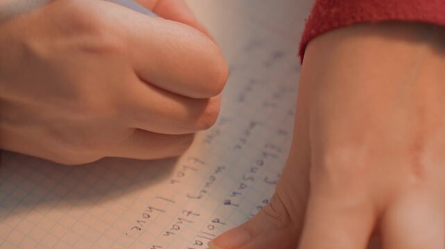 Close up of hands writing in blue ink on lined paper forming text. Red sleeve is visible next to hand. Notebook has grid pattern with handwritten content progressing steadily