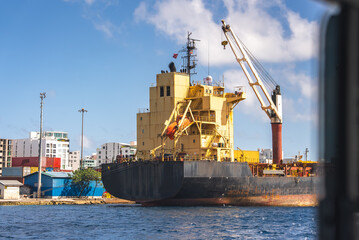 Fototapeta premium A large industrial cargo freighter ship rests docked at the port harbor in Male, Maldives under a partly cloudy sky near the urban coastline.