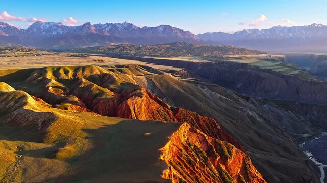 Aerial view of red canyon ridges illuminated by warm sunset light with distant snowy mountains in Anjihai Grand Canyon, Xinjiang, China