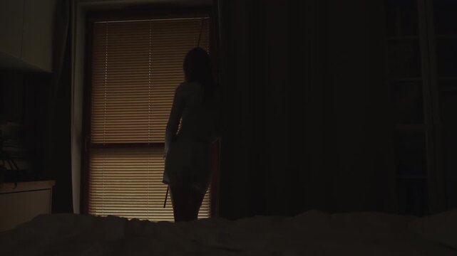 Woman standing by the window and covering the blinds then peeking outside. Suffering from depression and anxiety. Rear view