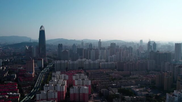 A wide aerial shot shows a dense modern urban city with high-rise buildings, roads, and distant hazy hills under a clear sky.
