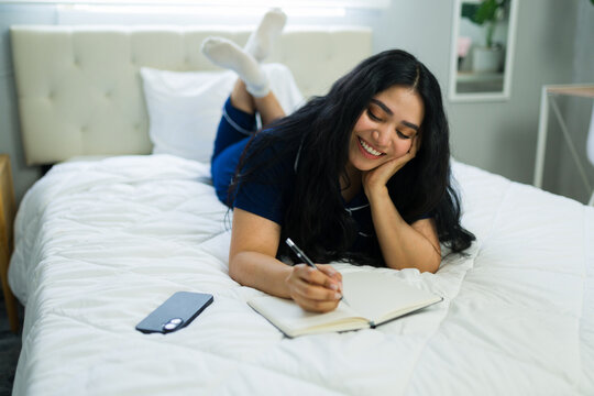 Happy woman writing in journal on bed, relaxing at home in pajamas, practicing self-care and mindfulness