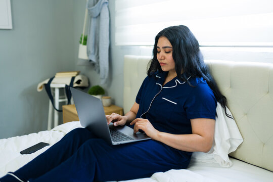 Young woman working from home in bed, typing on laptop while wearing pajamas in her bedroom