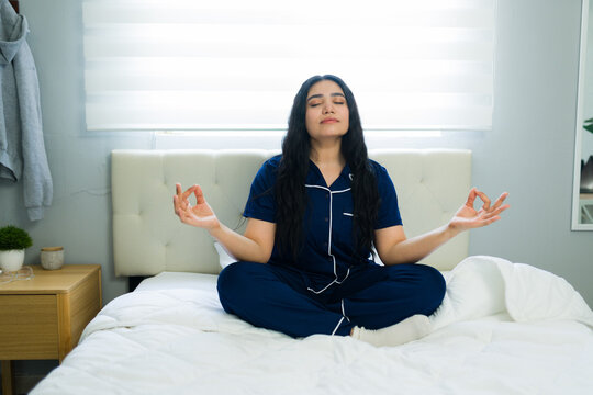 Relaxed woman in navy-blue pajamas sits up in bed, eyes closed, meditating to greet the morning with calm and mindfulness