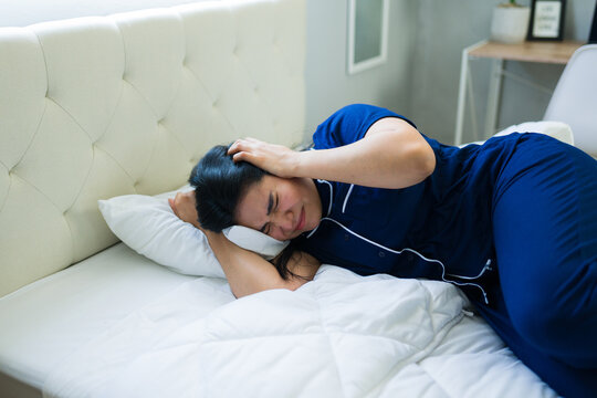 Woman suffering from severe headache or migraine in bedroom, feeling discomfort and stress, covering ears