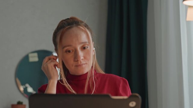 Young woman in red turtleneck is seated indoors interacting with open laptop. Woman adjusts earphones and looks down. Small potted plant appears briefly near her