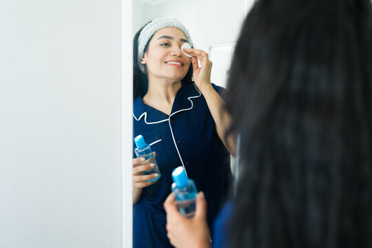 Hispanic woman standing in front of mirror, cleaning face with cleanser and cotton pad, wearing pajamas and headband