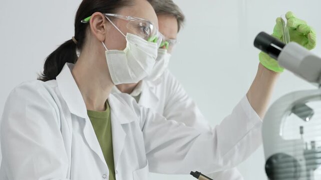 Two diverse scientists. Wearing lab coats. Safety goggles. And masks. Performing detailed research using a microscope in a clean laboratory setting