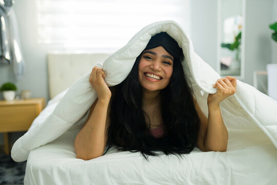 Happy young woman smiling from under duvet in bed, enjoying morning comfort and relaxation at home