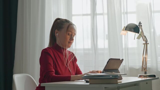 Subject types notes into digital reader from white table. Brass lamp and stacked books surround workspace area. Desk setup includes chair backrest and visible window view