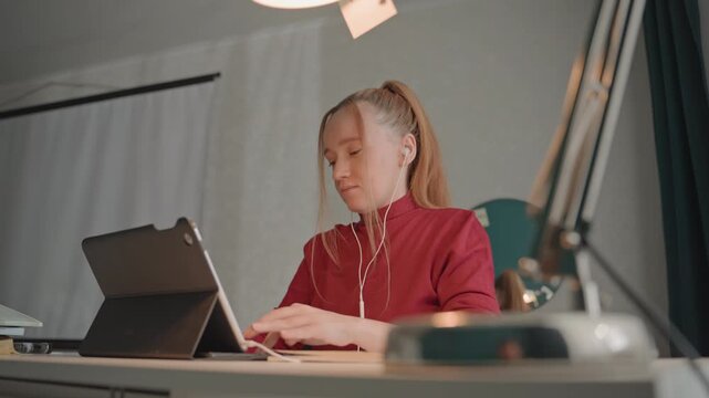 Young woman in red top works. Desk features open book closed laptop pen on white table. Background includes neutral walls hanging lamp partially featuring woman young red