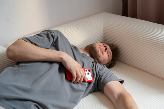 A man rests on a couch with a phone in his hand. His expression shows he is tired. The day is calm and relaxed