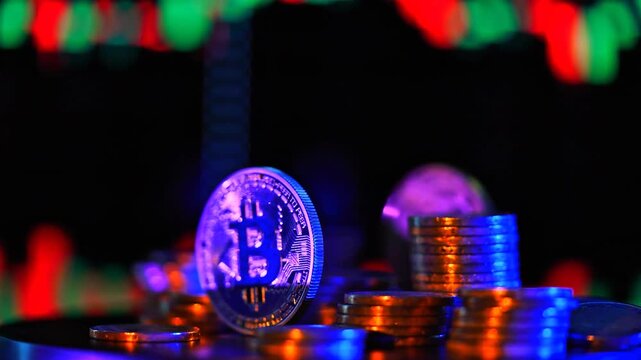 Golden Bitcoin crypto coin standing upright among stacks of coins, illuminated by dramatic blue and orange studio light against a blurry financial stock market candlestick chart.
