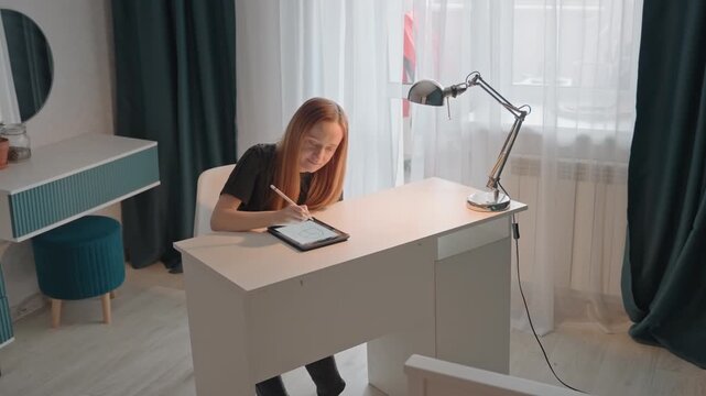 Young woman with long hair seated at white desk drawing. Desk features metallic lamp and teal ottoman nearby. Room has light wood flooring sheer curtains with woman young
