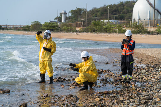 Environmental engineers in hazmat suits collecting and inspecting water samples near industrial gas storage tanks for pollution and safety monitoring.