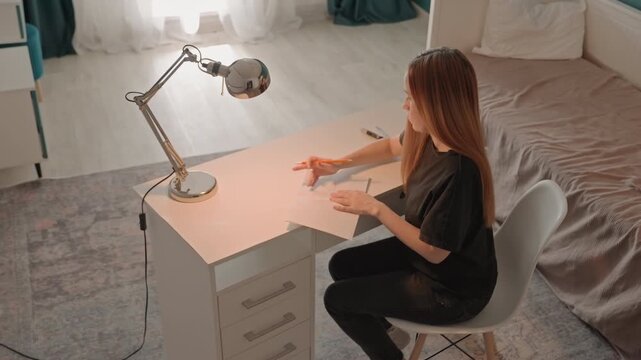 Young woman with long hair writes on paper at white desk. Woman sits upright as woman focuses on task involving drawing. Scene is well lit by natural light and artificial lamp