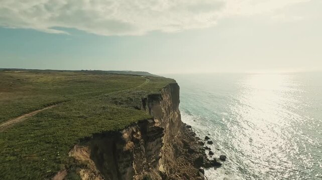 Aerial view captures cliff edge meeting ocean. Green grass blankets cliffside. Sunlight glints off calm sea waters. Sky shows soft clouds above. Wind rustles through grass gently