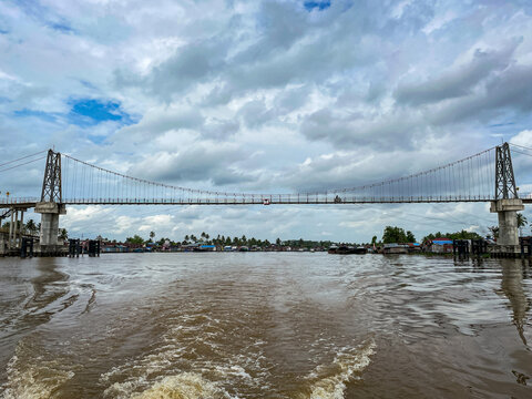 Wide suspension bridge spans murky river under cloudy sky. Boat's wake ripples across brown water beneath. Structural towers anchor bridge with taut cables.